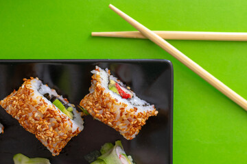 Top down view of a black square plate with multiple sushi rolls with sesame seeds and cucumber on green simple background with crossed chopsticks
