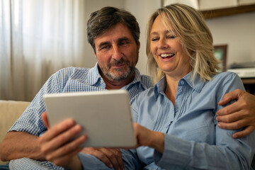 A happy mature couple is using a tablet for family entertainment while sitting on a sofa in living room at home.