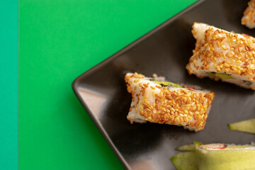 Top down close up view of sushi rolls with sesame seeds and cucumber on a black square plate. Flat on a bright green and light green background