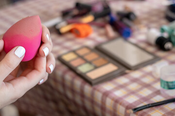 Fototapeta na wymiar Female hands with white fingernails holding a pink make up sponge. Daily routine of applying make up, accessories and brushes in the background