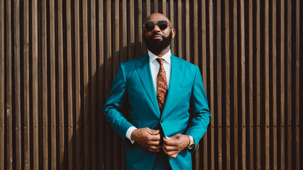 Fototapeta A handsome mature bald bearded African man in a sunglasses and a fashionable blue or teal costume with a tie is standing in front of a wall made of striped wooden timbers and fastening a suit button obraz