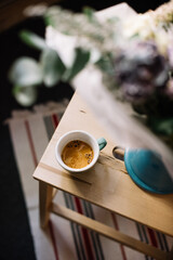 Delicious fresh morning cup of espresso coffee with a beautiful thick crema in a ceramic cup standing on the wooden coffee table with a flower bouquet blurred on the foreground, top view
