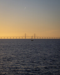 Fototapeta na wymiar A single sailing boat is out on the the calm ocean between Denmark and Sweden during sunset with the Oresund bridge in the background