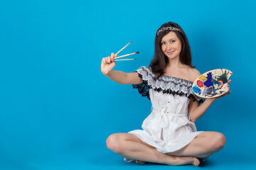 A beautiful girl with a brush and palette in her hands looks merrily at the camera. A model in a white dress poses on a blue background in the Studio. Copy space. Mock up