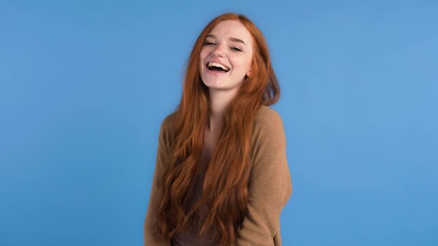 Red-haired model in brown t-shirt and cardigan. Clenched her fists and showing yes sign by hands , smiling while posing on blue background. Close up