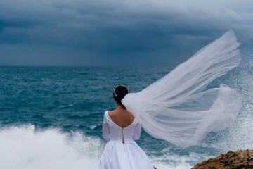 Fototapeta na wymiar Back view of brunette bride in white wedding dress and bridal veil on a cloudy day. Romantic beautiful bride posing near the sea with waves. The bridal veil flies in the wind