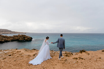 Fototapeta na wymiar Young wedding couple walking on a rocky beach near the blue sea. The groom holds the bride's hand. Beautiful wedding day
