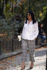 Fototapeta premium Autumn fashion outdoor. The girl in fashionable stylish white shirt , pants and sunglasses, autumnal lifestyle on the background of blurry yellow-green trees in the park. Vertical