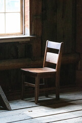 A singular wooden chair sitting on a wooden floor bathed in sunlight streaming through an adjacent window casting a peaceful glow of solitude.