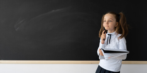Girl schoolgirl is at school. She is standing in front of a blackboard and is wearing a school uniform. In her hands is a notebook and she thought. Place for an inscription.
