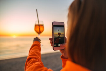 Fototapeta premium Girl Holding Glass of Drink on Beach at Beautyfull Romantic Sunset Taking Pictures with Phone. Glass and Hand Close Up. View From Back