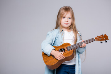 baby girl playing music on a small guitar. European girl with a guitar on a gray background. The concept of leisure and creativity