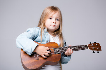Fototapeta premium baby girl playing music on a small guitar. European girl with a guitar on a gray background. The concept of leisure and creativity