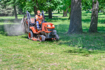Man mows the grass under trees in park by riding mowing machine in sunny day