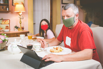 Father with his dauther are having breakfast in hotel with a protective mask and social distancing - Happy family having italian breakfast together in the kithen - The man is watching the tablet