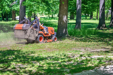 Man mows the grass under trees in park by riding mowing machine in sunny day