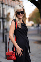 Fototapeta premium Autumn fashion outdoor. The blonde girl in fashionable stylish black overalls, sunglasses and red bag autumnal lifestyle on the background of blurry yellow-green trees in the park. Vertical
