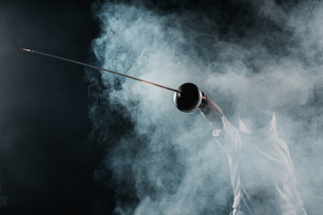 Low angle view of fencer holding rapier on black background with smoke