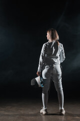 Back view of swordswoman holding fencing mask and rapier on black background with smoke