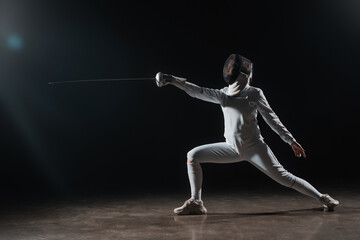 Fototapeta na wymiar Fencer in fencing mask doing lunge while training with rapier on black background