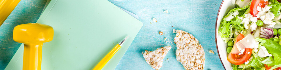 Healthy eating, slimming. dieting and weigh loss concept background. Fresh greenery summer salad with fruit, measuring tape, dumbbells and diet plan dairy notepad, flat lay above copy space