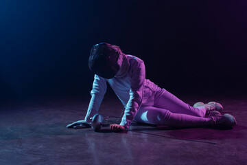 Swordswoman in fencing suit and mask holding rapier while sitting on floor on black background