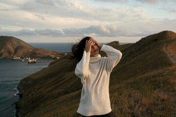 A girl in a white sweater with flowing hair touches her dreamy face against a picturesque landscape, hills and the sea