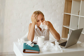 Young blond pretty sick with flu businesswoman sitting at her working place holding white napkins....