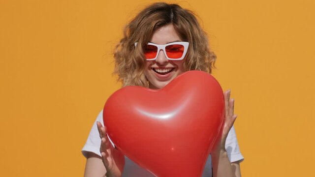 Portrait of happy woman with red heart balloon holding in her hands moving from side to side in front of her slow motion smiling camera yellow background summer. Emotions. Positive. Flirt. Valentine