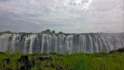 Unique Victoria Falls. The Zambezi River brings down numerous powerful streams of water through the edge of the gorge. In the foreground is another edge, covered with bright green grass. Cloudy.