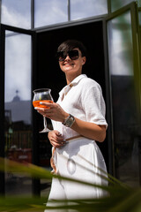 Beautiful woman with short brunette hair in white clothes and sunglasses. Fashion street photography. Fashion model posing with aperol cocktail on wood background.