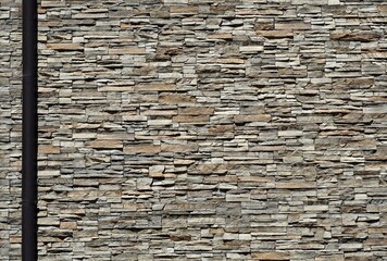 Stone cladding wall made of striped stacked slabs of natural brown and gray rocks with a black gutter onthe left. Panels for exterior 