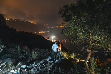 Two tourists on top of the mountain. Climbing, hiking trail. View of the night city. Romantic setting. Couple in love back view, look distance. Active lifestyle. The best view. Nerano, Italy