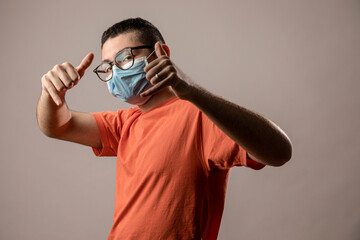 Ragazzo giovane con mascherina protettiva facciale, occhiali da vista neri e shirt arancione fa dei gesti da repper , isolato su sfondo grigio