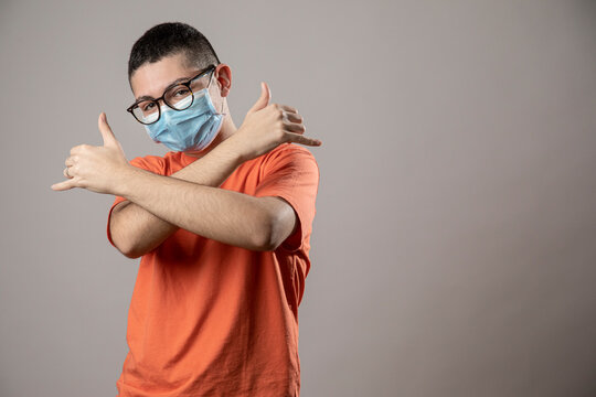 Ragazzo giovane con mascherina protettiva facciale, occhiali da vista neri e shirt arancione fa dei gesti da repper , isolato su sfondo grigio
