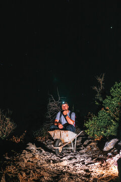 A Male Worker Is Sitting In A Wheelbarrow. Night View. Headlamp Illuminates The Trail. The Concept Of Heavy Utility And Construction Work In The Mountains On A Night Shift. Vertical Photo. Fun