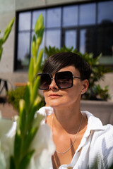 Beautiful woman with short brunette hair in white clothes and sunglasses. Fashion photography. Fashion model posing with flowers on wood background.