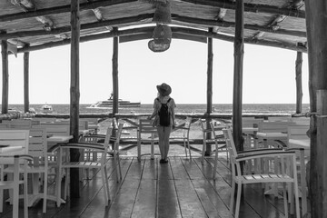 A girl stands alone in a wooden restaurant with sea views. Sunny summer day. Dressed in a hat and waterproof bag. Bay with boats and ships. Back view. Black and white. Nerano Napoli