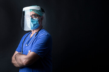 Fototapeta na wymiar medico in camice blu , cuffietta verde, visiera protettiva indossa una mascherina chirurgica incrocia le braccia isolato su sfondo nero