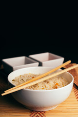 Chinese food bowl of rice with chopsticks and soy sauce and sweet and sour sauce on bamboo trivet and black background