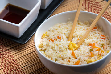Chinese food bowl of rice with chopsticks and soy sauce and sweet and sour sauce on bamboo trivet and black background