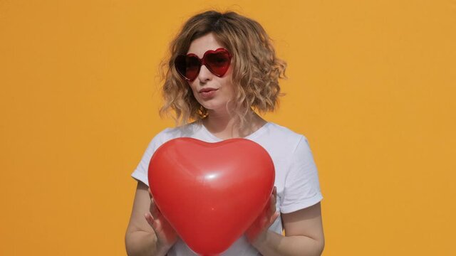 Portrait of happy woman with red heart balloon holding in her hands imitating how heart beats in front of herself smiling at camera on yellow background summer. Emotions Positive girl Flirt. Valentine