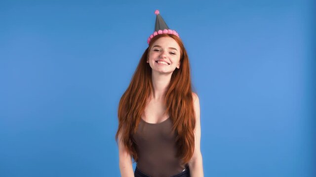 Ginger model in party hat and khaki shirt. She dancing, smiling and clapping hands while posing on blue background. Happy birthday concept. Close up, slow motion