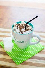 Coffee with white marshmallows, grated chocolate, a black straw in a mug with the inscription house, on a green napkin, on a wooden table.