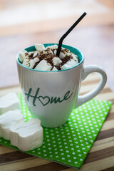 Coffee with white marshmallows, grated chocolate, a black straw in a mug with the inscription house, on a green napkin, on a wooden table.