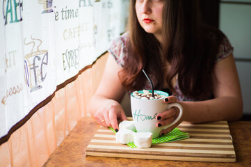 A young girl is waiting for a meeting in a cafe with a cup of coffee, with white marshmallow sprinkled with chocolate chips and a black straw on a wooden table.