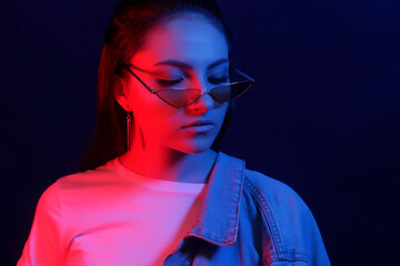 The young girl in a denim jacket and sunglasses bright neon blue and red light. The concept of beauty and fashion.