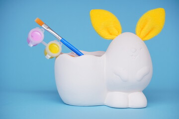 statuette of white figure of bunny or rabbit with yellow ears for decorating, Easter DIY with children. Multi-colored paints and brushes on blue background. Minimal easter concept