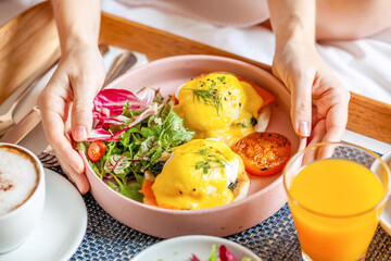 Breakfast in Bed Served with Cup of Coffee, Salad, Fresh Fruits and Eggs Benedict on Wooden Tray. Woman Hand Holding Plate with Fresh Food. Room Service in Hotel