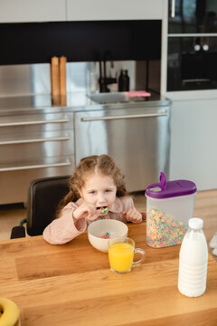 Cute Kid Eating Corn Flakes Near Glass Of Orange Juice And Bottle With Milk